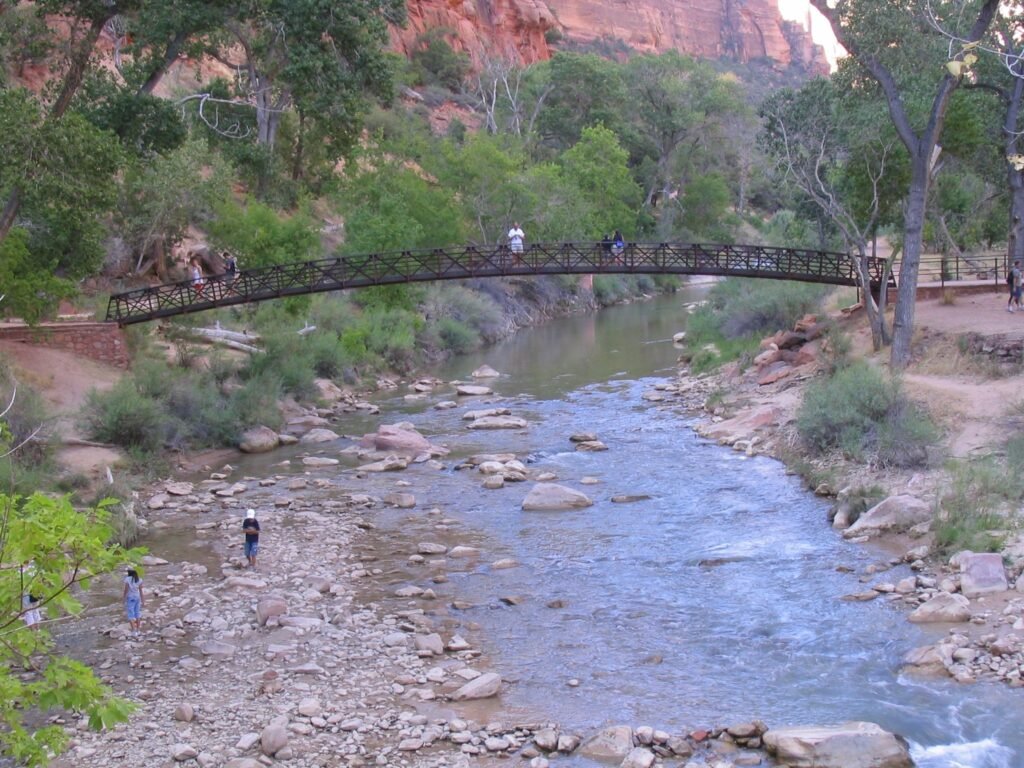 Families enjoying the Virgin River along the Riverside Walk in Zion, with kids cooling off in the shallow water beneath towering canyon walls on this flat, family‑friendly trail.
