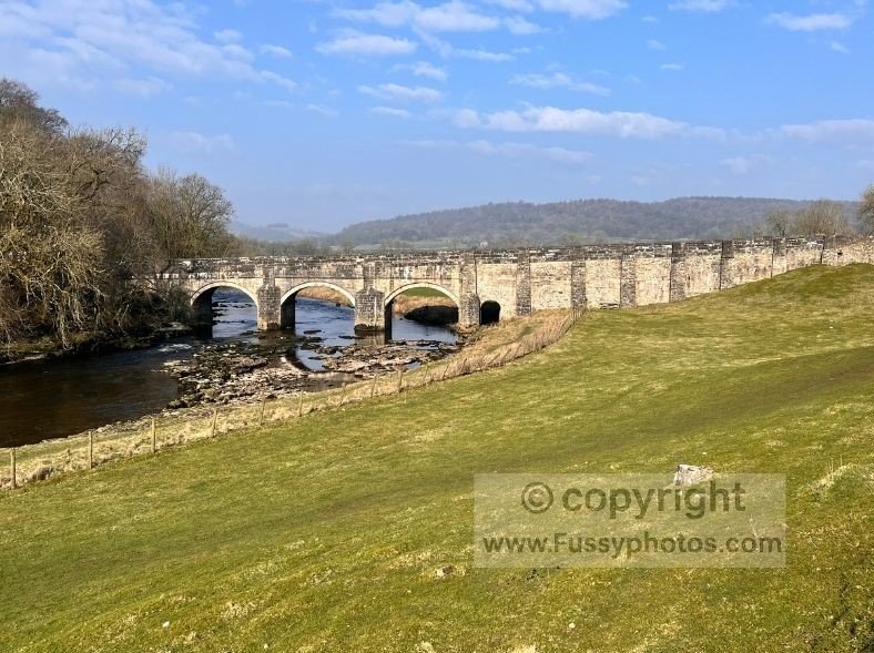 View looking west over the River Wharfe floodplain towards the Grade II‑listed Grassington road bridge at the start of the Dales Way.