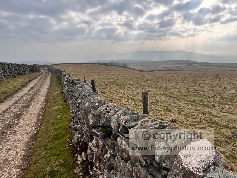 Path enclosed by high dry‑stone walls across open moorland, with distant sunlight piercing through the clouds near Yarnbury.