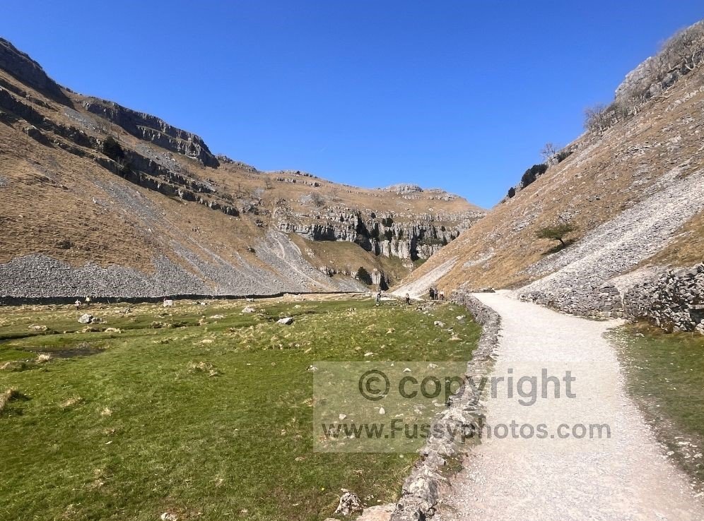 Flat riverside path inside Gordale Scar, showing the stream, steep limestone walls and the high viewpoint visible above the gorge.