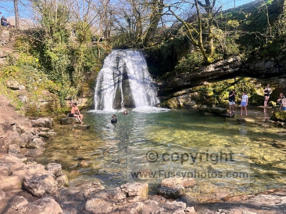 View of Janet’s Foss showing the waterfall, plunge pool and swimmers, with families gathered around the popular spot near Malham.