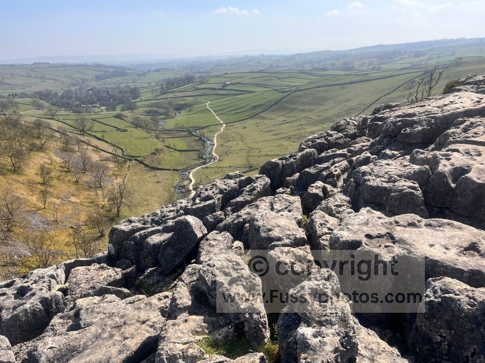 Limestone pavement above Malham Cove with a south‑facing view towards Malham, showing the Pennine Way on the classic village route running beside Malham Beck.