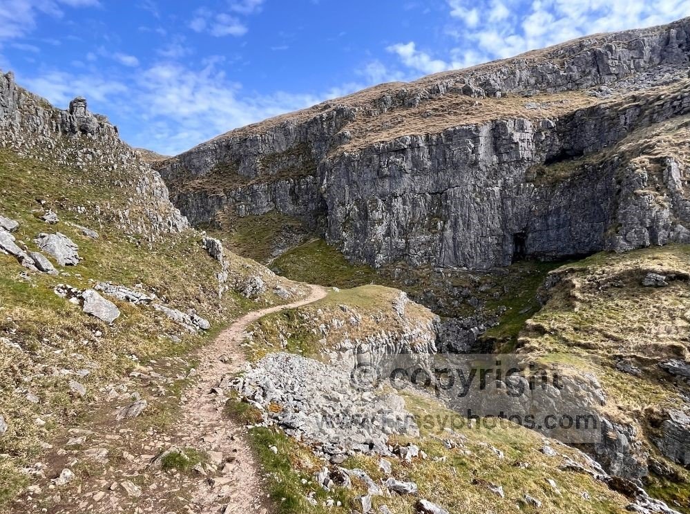 View of the Pennine Way north of Ing Scar, showing the high‑level path above the valley walls with caves and limestone formations on the ascent towards Malham Tarn.