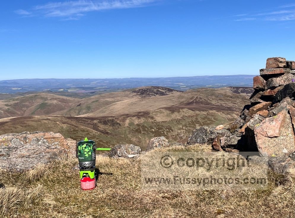Camping stove cooking a meal at Auchope Cairn, with scenic views behind.