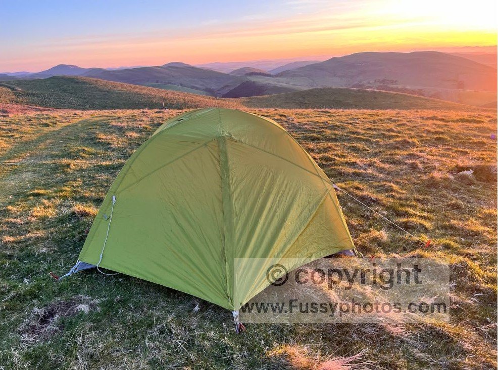 Tent on open ground beneath the Cheviots, framed by a bright sunset sky.