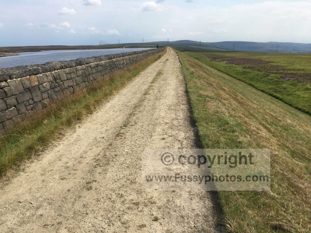 South‑facing view from the north end of Warland Reservoir, showing the long, straight moorland track and the same distant pylons seen earlier between Blackstone Edge and White Holme Reservoir.