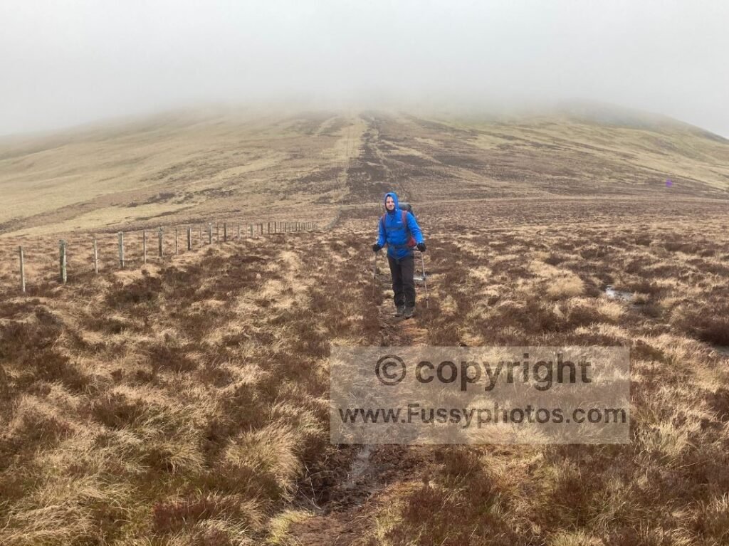 South‑facing view towards The Schil, hidden in cloud, with a soaked walker in full rain gear standing in the foreground.