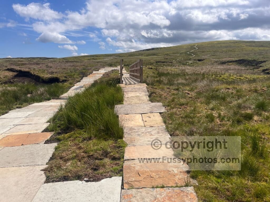 View uphill to Nine Standards Rigg from the boggy stretch featuring both a wooden bridge and neighbouring paving stones, taken partway through the long walk out to Kirkby Stephen.