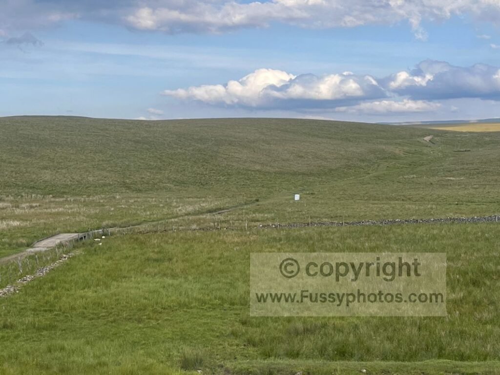 Large open moorland with a white fridge honesty box in the centre of the scene, set among sheep‑grazed fields on the approach to Sunbiggin Tarn.