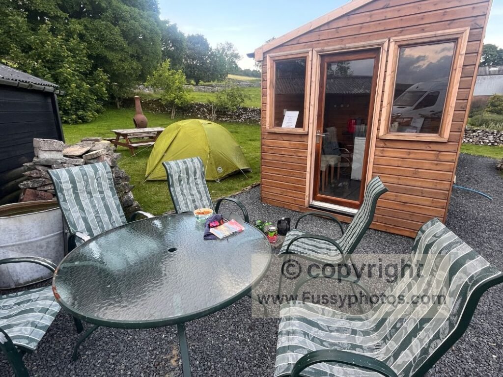 Small tent set up next to the honesty‑box shed at Sunbiggin Farm, with chairs and a table visible outside in an otherwise empty moorland setting.