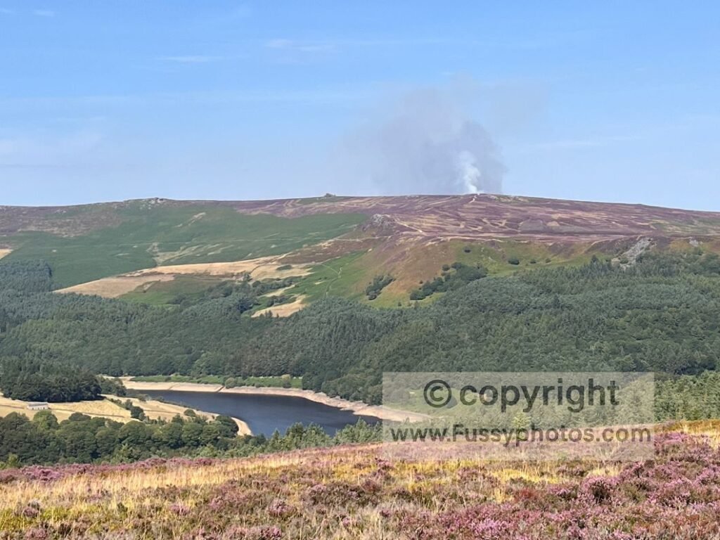 Summit panorama from Win Hill over Ladybower Reservoir with wildfire smoke on the Derwent Moors; long‑distance view to the north‑east showing reservoir water, surrounding ridges, and haze from a moorland fire.