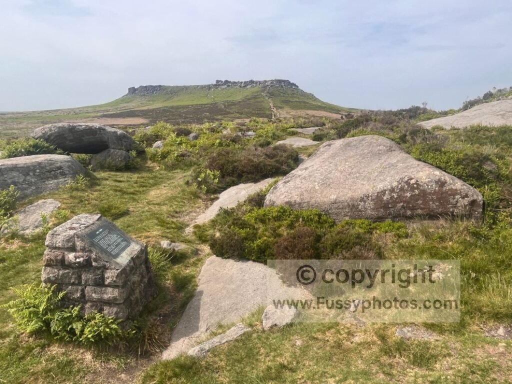 Northward view from Carl Wark, showing rocks and the sign in the foreground with Higger Tor visible in the distance.