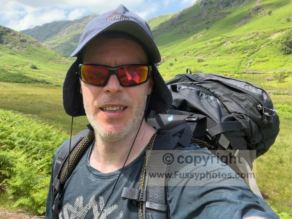 Walker cooling a t‑shirt in a Lake District beck during a hot, humid Coast to Coast day to manage heat and fatigue.