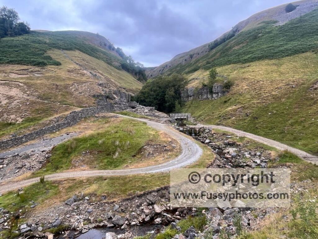 Swaledale low‑route bridge — wooden bridge and small waterfall on the low‑level Coast to Coast with high‑level terrain above.

