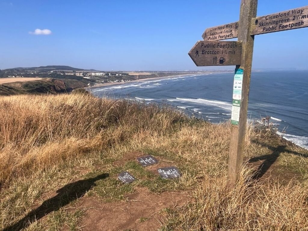 Cleveland Way trail sign on Hunt Cliff overlooking Saltburn‑by‑the‑Sea, with mental‑health support signs placed along the grassy cliff edge.