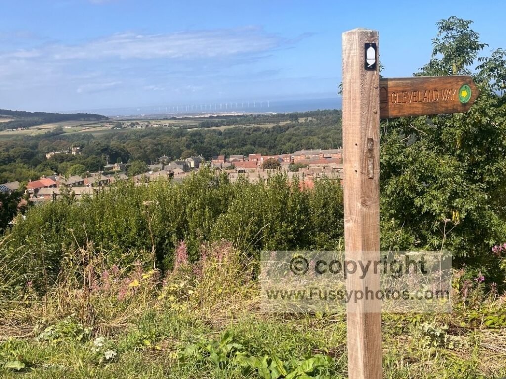 Cleveland Way trail sign at the southern end of Skelton, with coastal views and an offshore wind farm visible from the elevated viewpoint.