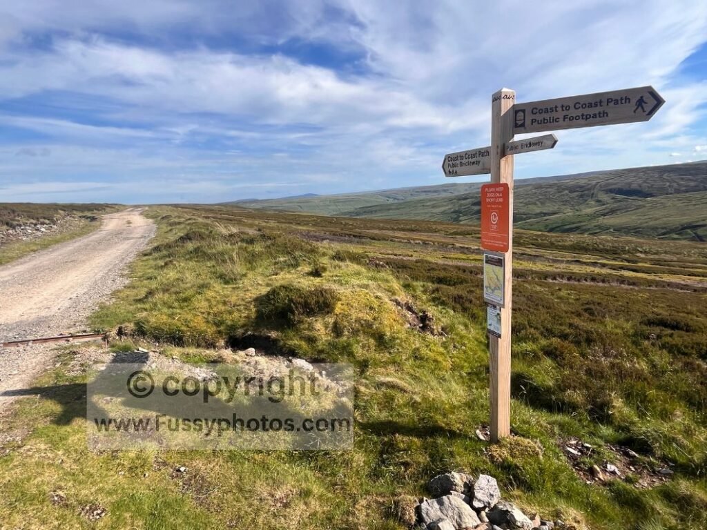  Two‑finger signpost above Gunnerside Gill in Swaledale on the Coast to Coast, showing direction choices for east → west and west → east walkers.