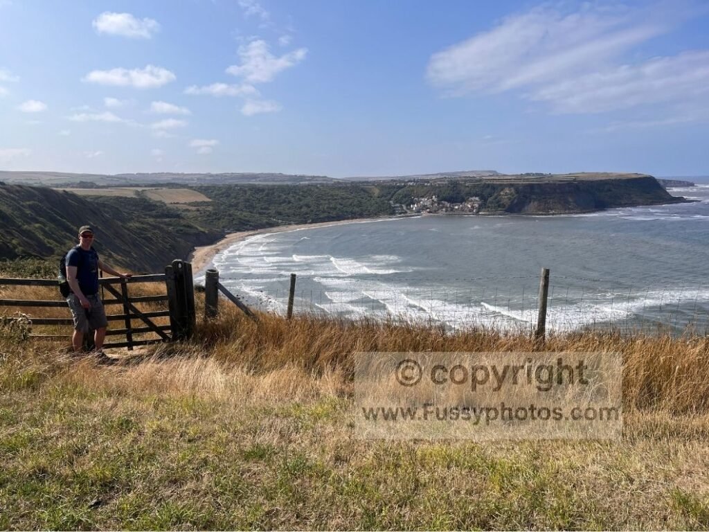 High‑cliff view towards Runswick Bay from the Cleveland Way, with the walker posing beside a gate above Hill Stones.